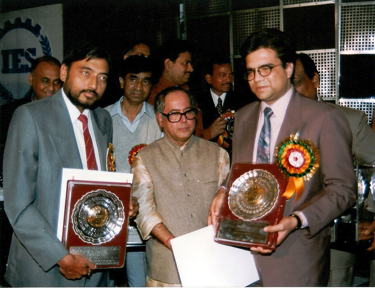 Two men of the company receiving the Export Excellence Award in February 1997, each holding a shield, with the presenting official between them.